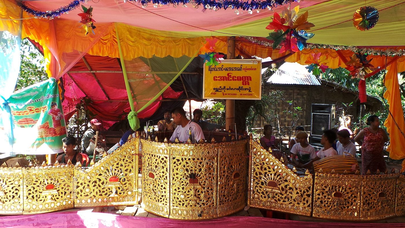 Ein Hsaing-waing-Ensemble spielt hinter seinen vergoldeten, geschnitzten Rahmen unter einem bunten Pandal bei einer Dorfzeremonie